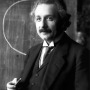 Albert Einstein in a suit and tie standing in front of a chalkboard 